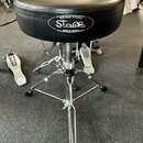 Černý bubeníckýstoliček s polstrovaným sedadlem z umělé kůže a chromovanou třínohou konstrukcí. Na boku sedadla je bílý nápis DRUMS STAND Stable MADE…