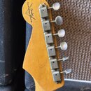 Záběr na zadní stranu hlavy kytary z přírodního dřeva. V horní části je černý nápis "Custom Shop" a logo "Fender". Na pravé straně jsou osazeny šest…