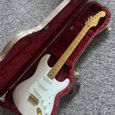 Bílá elektrická kytara typu Stratocaster s javorovým hmatníkem a zlatým hardwarem v otevřeném hnědém tvrdém kufru s červeným polstrováním. Na hlavě k…