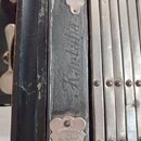 Detailní pohled na část hudebního nástroje s kovovými lištami a černým povrchem. Na látkovém pásu je nápis „Kantalia“. Dole se nachází kovový štítek…