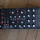Černý semi-modulární analogový polyrytmický syntetizér s nápisy SUBHARMONICON a moog. Přední panel obsahuje množství stříbrných otočných ovladačů, če…