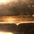 Detailní záběr na štítek uvnitř hudebního nástroje. Na štítku je černým textem nápis: Jacobus Stainer ex Absam prope Oenipontum fecit Cremona. Papír…
