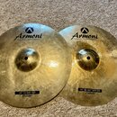Dvě mosazné činely s viditelným rýhováním povrchu. Oba mají v horní části logo a nápis Armoni HANDMADE TURKISH CYMBALS. Levý činel má dole černý štít…