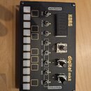 Černý elektronický modul s texturovaným povrchem a zlatými nápisy. Na zařízení jsou bílé tlačítka, několik přepínačů a dva černé otočné knoflíky. Jso…