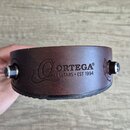 Hnědý kožený řemen s černým gumovým podkladem. Na povrchu je vyražené logo a nápis ORTEGA® GUITARS • EST 1994. Řemen je vybaven dvěma kovovými šroubo…