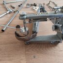 Fotografie zobrazuje kovový mechanismus pedálu, pravděpodobně pro bicí nástroj, v šedé barvě. Součástí je pružina a hnědý kožený pásek, který vykazuj…