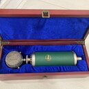 Otevřený dřevěný kufřík s modrým sametovým vnitřkem, ve kterém leží kondenzátorový mikrofon. Mikrofon má stříbrnou síťovou mřížku a zelený válcový ko…