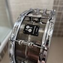 Detailní pohled na chromovaný obruč a korpus bubnu. Na černé čtvercovédesce je bílé logo dvou kruhů a nápisy KOMPRESSOR a SONOR. Na chromovaném obruč…