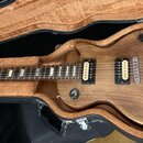Elektrická kytara s hnědým dřevěným povrchem v černém tvrdém kufru s hnědým polstrováním. Nástroj má dva humbuckery, čtyři oválné ovládací knofíky a…