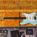 Světle modrá elektrická kytara typu Stratocaster v tvrdém kufru s žlutým polstrováním. Na hlavě hmatníku je nápis Fender a na krytce snímačů jsou vid…