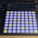 Černý MIDI kontrolér s mřížkou podsvícených čtvercových padů v bílé a zelené barvě. V horní části je zapnutý displej s texty Utility, Happy Bass, Osc…