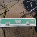 Zelený transportní kufr s hliníkovými hranami a vrchními zámky. V pravém horním rohu je nalepený štítek s nápisem KOSMI BOARDS. Povrch kufrů vykazuje…