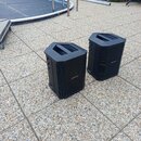 Dva černé plastové reproduktory s hranatým tvarem a prohlubní v horní části. Na přední straně obou kusů je umístěno stříbrné logo BOSE. Povrch je mat…