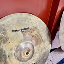 Detailní pohled na mosazný činel s viditelným opotřebením povrchu a skvrnami. Na povrchu jsou nápisy „HiHat Bottom“ a „14"/36 cm“ a v dolní části log…