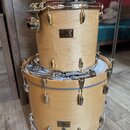 Fotografie zobrazuje tři dřevěné bubny v přirozené světlé barvě s kovovým hardwarem. Na dvou z nich je viditelný nápis Pearl Masters Custom. Povrch n…
