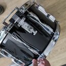 Černý buben s chromovaným obručem a mechanismy. Na korpusu jsou vidět čtyři chromované držáky s vlnitým tvarem a centrální mechanismus s pružinou. Po…