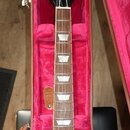 Elektrická kytara v hnědém sunburst provedení umístěná v červeném tvrdém kufru. Na hlavě hmatníku je bílý nápis Les Paul a na vnitřní straně víka kuf…