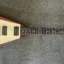 Elektrická kytara s V-tvarým korpusem v červenohnědé barvě a krémovými plastovými kryty. Na hlavě hmatníku je bílý nápis Gibson. Nástroj má dva humbu…