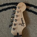 Fotografie zachycuje hlavu elektrické kytary z jasného dřeva s chromovanými ladičkami. Na povrchu je černý nápis Squier BY FENDER a STRATOCASTER®. V…