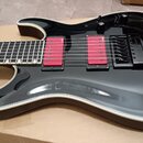 Černá elektrická kytara s bílým lemováním korpusu a dvěma červenými humbuckery. Na snímačích je viditelný nápis "ESS". Kytara má černý most s nápisem…