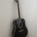 Černá akustická kytara s bílým lemováním korpusu a hnědým hmatníkem. Na hlavě je bílý nápis Epiphone. Nástroj je opřen o světlou stěnu a stojí na bíl…