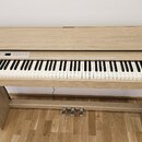 Digitální piano v dřevěném provedení světlé barvy s integrovaným stojanem a třemi pedály. Na pravém okrave korpusu je viditelné logo Roland. Ovládací…