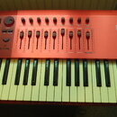 Červený MIDI kontrolér s klávesnicí. Na pravé straně je bílé logo DX6 a nápis Intelligent Keyboard Controller. Ovládací panel obsahuje několik posuvn…