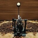 Černý bubenový pedál s kovovými prvky a bílým plátýčkem šlapáku. Na kovové části mechanismu je svisle napsán nápis "TAMA". Povrch pedálu vykazuje mír…
