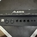 Černý elektronický modul značky ALESIS s řadou vstupů a výstupů. Na horní části jsou bílé nápisy USB MIDI, OUT, MIDI, IN, TOM 4, CRASH 2, R OUTPUTS L…
