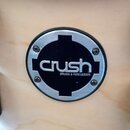 Detailní pohled na kulatý kovový emblem přišroubovaný k dřevěnému povrchu. Černý střed s bílým nápisem crush a menším textem DRUMS & PERCUSSION je ob…