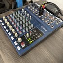 Modrý mixážní pult s nápisem YAMAHA MIXING CONSOLE MG10/2. Zařízení obsahuje řadu regulačních knoflíků v bílé, modré a červené barvě a několik posuvn…