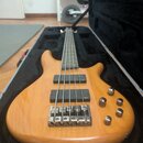 Elektrická basová kytara s přírodním dřevěným povrchem v černém tvrdém kufru. Nástroj má čtyři struny, dva černé humbuckery a čtyři ovládací knoflíky…