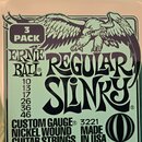 Fotografie znázorňuje balení kytarových strun v odstínu zlaté a bílé barvy. Na obalu je nápis ERNIE BALL REGULAR SLINKY, grafika orla a označení 3 PA…