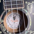 Černý ukulele s bílými grafickými prvky ve tvaru stylizovaných postav. Uvnitř ozvučovací otvory je bílá štítková nálepka s logem Fender a nápisy BILL…