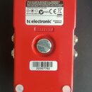 Spodní strana červeného efekt pedálu z kovu. Na povrchu jsou dvě nálepky s nápisy tc electronic, DESIGNED IN DENMARK, BATTERY?, 9V DC/100mA, NIB39 a…