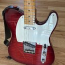 Elektrická kytara s červeným průsvitným lakem a viditnou strukturou dřeva. Nástroj má bílý pickguard, javorový hmatník a chromované kovové části. Na…