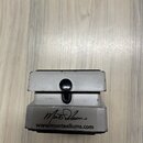 Šedý kouskový efekt s kovovým povrchem. Na spodní části je černý podpis a nápis www.monteallums.com. Povrch vykazuje drobné oděrky a známky používání.