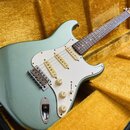 Světle zelená elektrická kytara typu Stratocaster s bílým pickguardem a krčkem z tmavého dřeva. Nástroj leží v otevřeném kufru s žlutým polstrováním.…