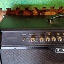 Černý kytarový kombozesilovač s nápisem SPIDER IV a logem LINE 6. Na ovládacím panelu jsou stříbrné ovratidla s popisky DRIVE, BASS, MID, TREBLE a CH…