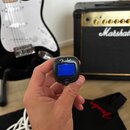 Ruka drží malý černý ladič s modrým displejem, na kterém je nápis Fender a označení FT-1. V pozadí je vidět černá elektrická kytara s bílým pickguard…