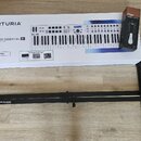 Na fotografii je krabice s nápisy ARTURIA a KEYLAB ESSENTIAL 61, na které je zobrazen bílý MIDI kontrolér. Vedle krabice stojí úzký černý obal značky…