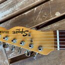 Detail hlavy elektrické kytary z dřeva s viditnou masivním žilováním. Na povrchu jsou nápisy Squier, "Fender" a TELECASTER. Kytara má šest chromovaný…