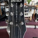 Černý hlavník elektrické kytary s bílým lemováním. V horní části je bílý nápis GRETSCH a svisle pod ním nápis ELECTROMATIC. Jsou vidět šest kovových…