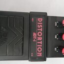 Černý kytarový efekt pedál s nápisy DISTORTION a ADT-1 v červené barvě. Na levé části je vyražen nápis ARIA a dekorativní vlnité linky. Pravá část ob…