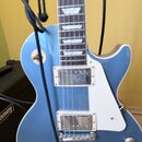 Modrá elektrická kytara s tvarem singlecut a bílým pickguardem. Nástroj má dva humbuckery s chromovanými kryty a čtyři ovládací knoflíky. V pozadí je…