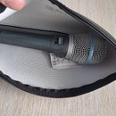 Černý dynamický mikrofon s kovovou síťkou a modrým prstencem, umístěný v černém zipovém pouzdře s bílou výstuhou. Na těle mikrofonu je vidět nápis "S…