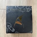 Černá čtvercová krabice s nápisem SIMPLIFIER DLX a textem ZERO WATT DUAL CHANNEL & REVERB STEREO AMPLIFIER. Uprostřed je zobrazen zlatý stylizovaný s…