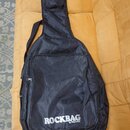Černý polstrovaný obal na hudební nástroj s ramenním popruhem. Na přední straně je bílý nápis ROCKBAG a pod ním menší text by Warwick. Obal má dvě zi…