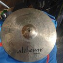 Kruhový činely z kovu s viditelným rýhováním povrchu. Na spodní části je černý nápis „alchemy cymbals“. V horní části je kurzívní nápis „Professional…