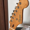 Detail hlavy dřevěné elektrické kytary v přirozené barvě. Na dřevě je černý nápis Squier BY FENDER a STRATOCASTER®. Jsou vidět kovové ladičky a strun…